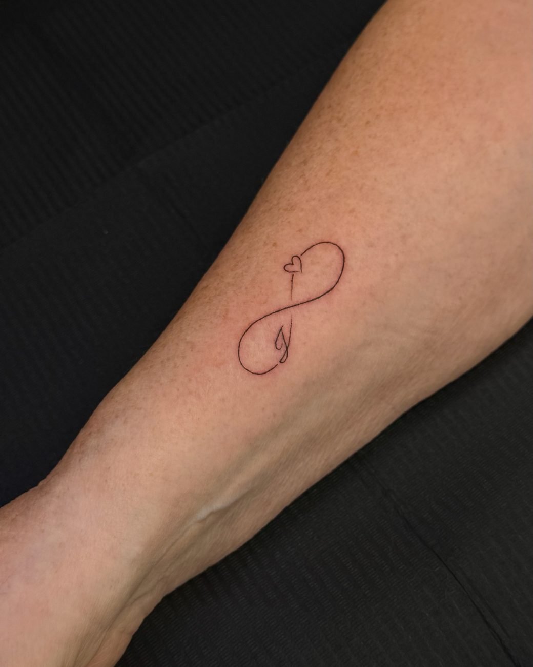 Fine line-tatuering av ett oändlighetstecken och ett hjärta på underarmen
